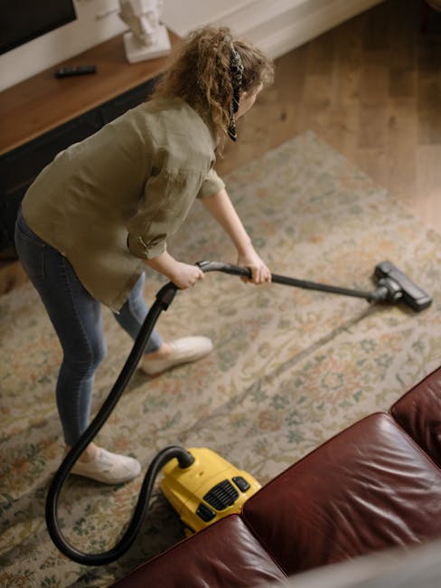 A woman engaged in surface cleaning within a living room, using a yellow vacuum cleaner with a long black hose attachment to thoroughly clean a patterned area rug. The room features a wooden floor adjacent to the rug, and a dark red leather sofa is partially visible in the foreground. She is wearing casual clothing, including a beige jacket and blue jeans, with her hair tied back. The background shows a wooden TV stand, a small decorative item, and neutral-colored walls. The scene is well-lit with natural light, emphasizing the cleanliness and maintenance of the living space, in line with domestic cleaning and deep cleaning processes promoted by Carpet Cleaning Catford.