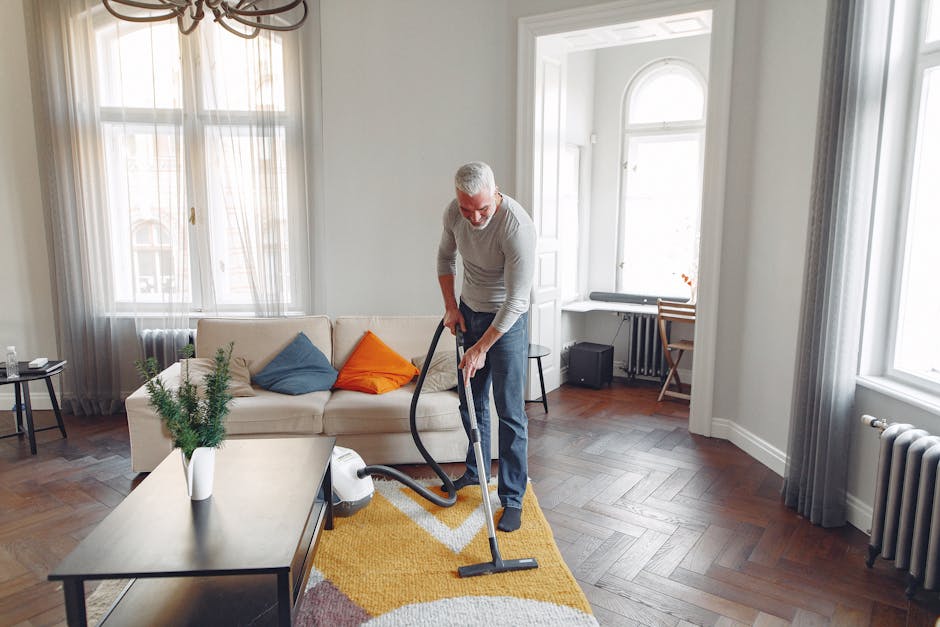 A woman engaged in surface cleaning within a living room, using a yellow vacuum cleaner with a long black hose attachment to thoroughly clean a patterned area rug. The room features a wooden floor adjacent to the rug, and a dark red leather sofa is partially visible in the foreground. She is wearing casual clothing, including a beige jacket and blue jeans, with her hair tied back. The background shows a wooden TV stand, a small decorative item, and neutral-colored walls. The scene is well-lit with natural light, emphasizing the cleanliness and maintenance of the living space, in line with domestic cleaning and deep cleaning processes promoted by Carpet Cleaning Catford.