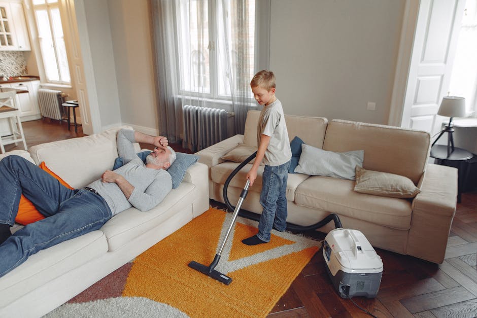 A young boy using a vacuum cleaner to clean a light-colored carpet in a living room, while an older man lies on a sofa, resting with a hand on his forehead. The room features large windows with sheer curtains, wooden flooring, and a beige sofa with pillows. Cleaning equipment, including a portable vacuum, is visible, and the space appears clean and well-maintained. This scene exemplifies domestic cleaning and surface maintenance, with natural lighting highlighting the tidy and hygienic environment. Carpet Cleaning Catford provides thorough cleaning services to ensure homes like this are hygienic and inviting.