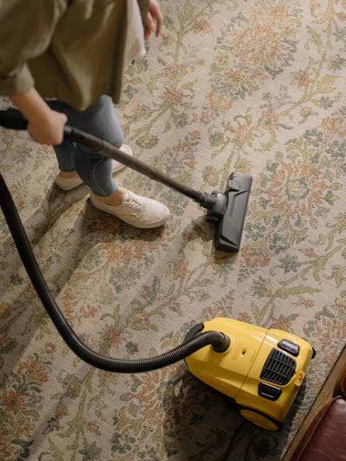 A person kneeling on a patterned carpet in a domestic setting, performing surface cleaning with a yellow portable vacuum cleaner. The individual, dressed in a beige jacket and jeans, is focused on vacuuming the area, which appears clean and well-maintained. The vacuum cleaner has a black hose and a reflective mirror inside its open top compartment, indicating active use. Natural lighting illuminates the space, highlighting the detailed floral design of the carpet. This image reflects the professional cleaning services offered by Carpet Cleaning Catford, emphasizing deep cleaning and hygiene maintenance suitable for home environments.