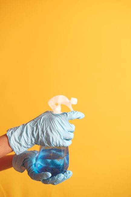 A hand wearing a light blue disposable glove is holding a transparent spray bottle containing a clear cleaning solution, with the nozzle directed upward. The background features a plain, vibrant yellow wall, and the lighting is bright and even, highlighting the cleanliness and sterile appearance of the cleaning item. This image emphasizes surface cleaning and sanitisation processes as part of domestic cleaning services offered by Carpet Cleaning Catford, aligned with deep cleaning advice for Rushey Green flats in SE6. The focus is on hygiene and professional cleaning tools suitable for residential environments.