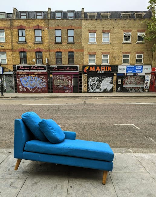A bright blue chaise lounge with two matching cushions is placed on the sidewalk in front of a row of commercial shopfronts with graffiti-covered metal shutters, situated along a bustling city street 