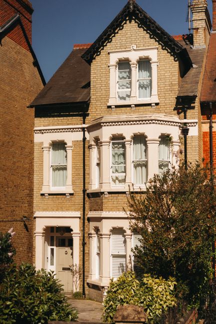 A three-story Victorian-style house with yellow brickwork, white decorative window frames, and a slate roof, situated in a residential area with neighboring brick buildings. The front garden features lush greenery and small bushes, with sunlight casting shadows on the facade, highlighting the architectural details. This image is related to residential cleaning services provided by Carpet Cleaning Catford, specializing in surface and deep cleaning for homes within the Catford SE6 area, including end of tenancy cleaning for flats on Brownhill Road.