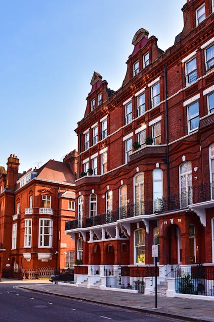 Photograph of a red-brick residential building on Brownhill Road in Catford SE6, featuring multiple stories with large sash windows, decorative brickwork, and small balconies with ornate black iron railings. The building’s concrete steps lead up to an entrance with white trim, and the facade appears clean and well-maintained under bright daylight, with a clear blue sky background. The street in front is quiet, with a lamppost and minimal traffic, reflecting a tidy urban setting. This image illustrates the exterior appearance of the property prior to or after professional cleaning services by Carpet Cleaning Catford, which specializes in domestic and commercial surface cleaning, deep cleaning, and sanitisation of residential buildings.