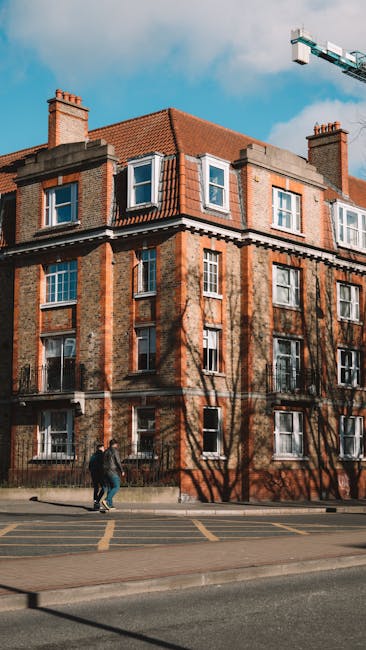 A three-story Victorian-style house with yellow brickwork, white decorative window frames, and a slate roof, situated in a residential area with neighboring brick buildings. The front garden features lush greenery and small bushes, with sunlight casting shadows on the facade, highlighting the architectural details. This image is related to residential cleaning services provided by Carpet Cleaning Catford, specializing in surface and deep cleaning for homes within the Catford SE6 area, including end of tenancy cleaning for flats on Brownhill Road.
