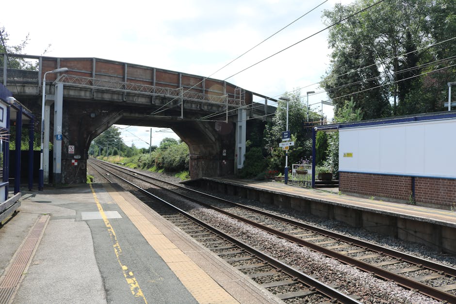 An outdoor railway station platform adjacent to railway tracks, featuring concrete flooring with tactile paving in yellow and grey, a blue metal shelter on the left side, and a white brick building on the right. Overhead, a steel bridge spans across the tracks, supported by brick and metal structures, with visible electrical lines running above the tracks. The area appears clean and well-maintained, with surrounding greenery and trees in the background, under a partly cloudy sky. This setting highlights the importance of surface cleanliness and safety in public transportation environments, aligning with deep cleaning and sanitisation practices promoted by Carpet Cleaning Catford for maintaining hygiene at commuter stations near Catford Bridge.
