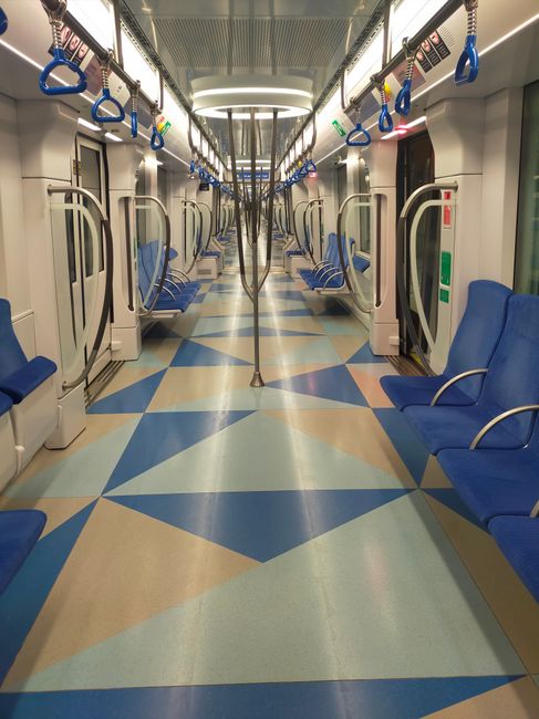 Interior view of a clean, empty train carriage with patterned fabric seats in shades of blue and purple, arranged in rows along both sides of the aisle. The white plastic walls and ceiling are spotless, with bright overhead lighting providing even illumination. Blue vertical and horizontal handrails are securely fixed along the sides and above the seats, assisting passengers during travel. The floor, made of dark textured material, appears well-maintained and free of debris. The windows show reflections of the interior lighting, emphasizing the cleanliness and hygiene of the space. This setting illustrates professional surface cleaning and maintenance, consistent with the services offered by Carpet Cleaning Catford for commercial environments such as public transportation.